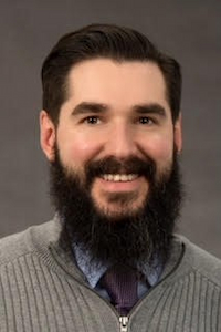 A light skin toned man with a brown beard, mustache, and hair. He wears a blue button down, navy tie, and a grey zip up cardigan.