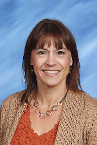 A photo of a woman with brown hair and light skin wearing an orange shirt, and a beige sweater. She is smiling.