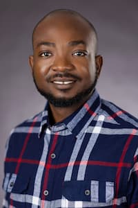 A man with dark skin, black hair, beard, and mustache. Wearing a flannel button up shirt and smiling.