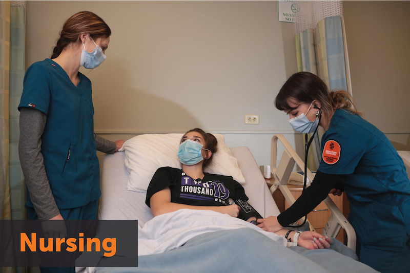 Two women in scrubs and masks stand around a patient on a hospital bed. Text: Nursing. Image links to program homepage.
