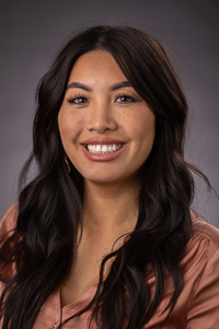 A woman with medium dark skin tone and black hair. She is wearing a light brown shirt.