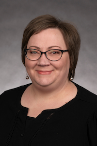 A light skin toned woman with short brown hair. She is wearing glasses, pearl earrings, and a black shirt under a black cardigan. She is smiling.