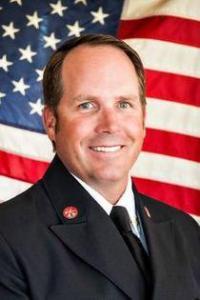 A man with medium skin tone and brown hair wearing a suit and standing in front of a United States flag