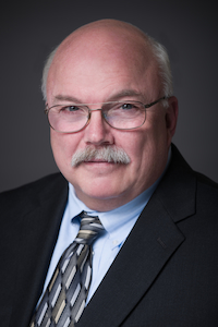 A man with light skin tone with balding white hair and a white mustache. He is wearing a light blue button down, a grey tie, and a black suit jacket.