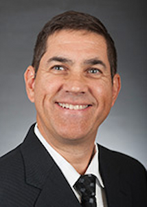 A photo of a man with medium skin tone wearing a white shirt with a black tie and a black suit jacket. He is smiling
