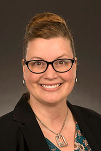 A light skin toned woman with black glasses and brown hair in a bun. She is wearing a blue shirt with a black blazer and a silver chain necklace, she is smiling.