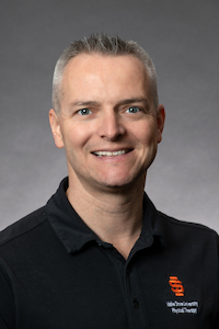 A man with light skin and short grey hair wearing a black polo shirt.