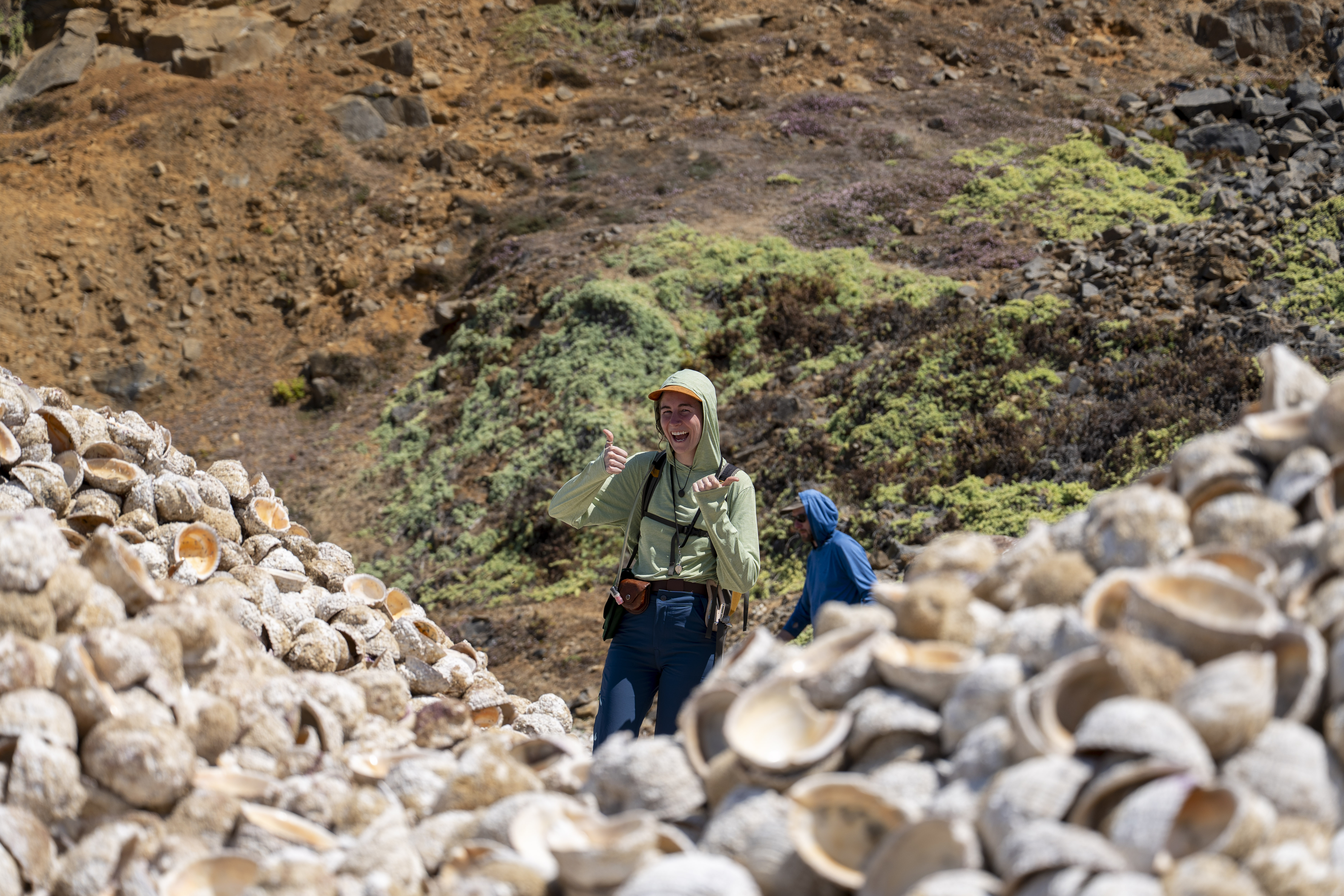 A smiling geologist in a green sun hoodie gives a thumbs-up while standing between massive mounds of seashells in a rugged, sun-drenched landscape.