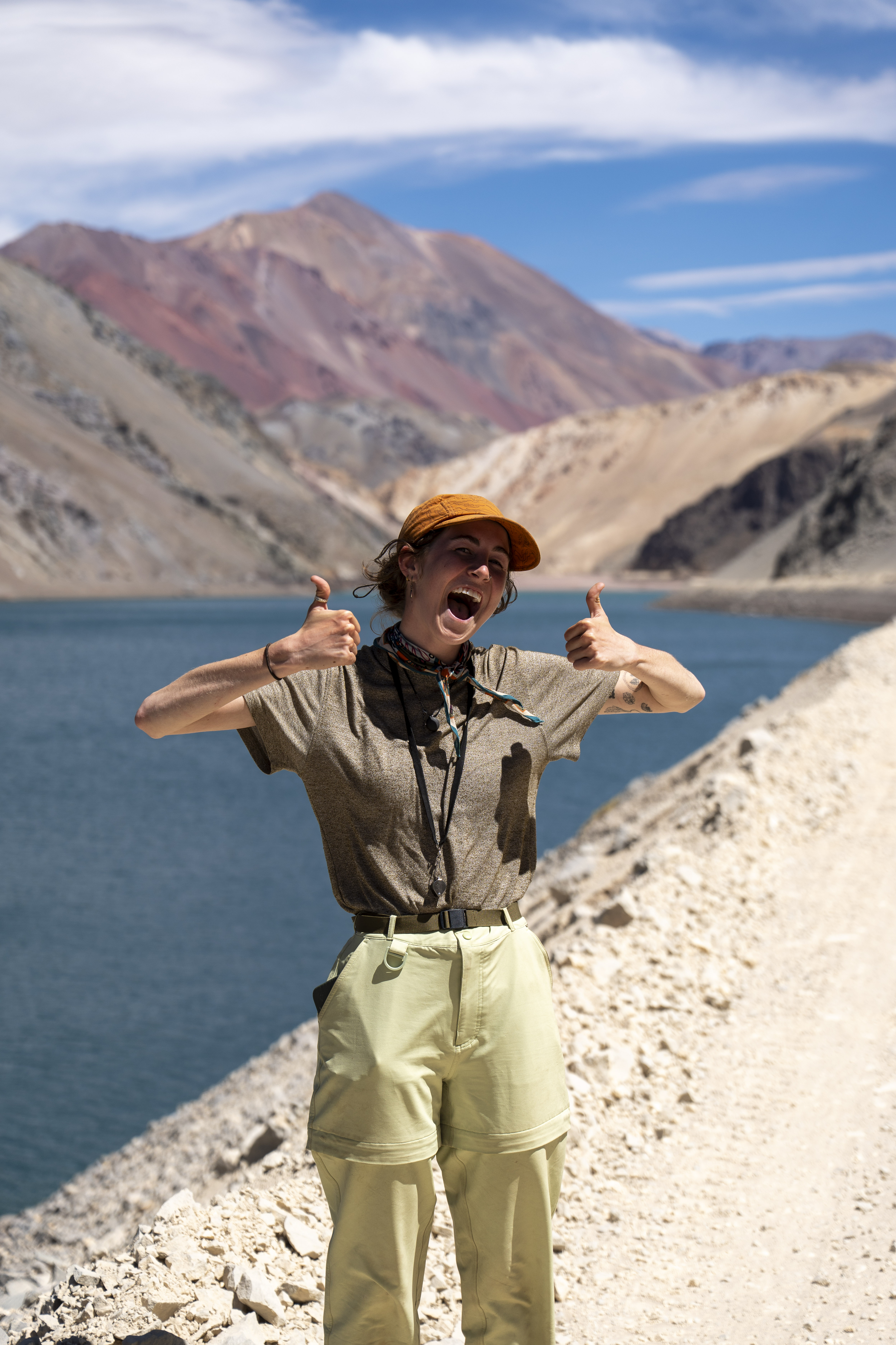 A cheerful geologist in an orange cap gives a double thumbs-up in front of vibrant blue waters and reddish mountains.