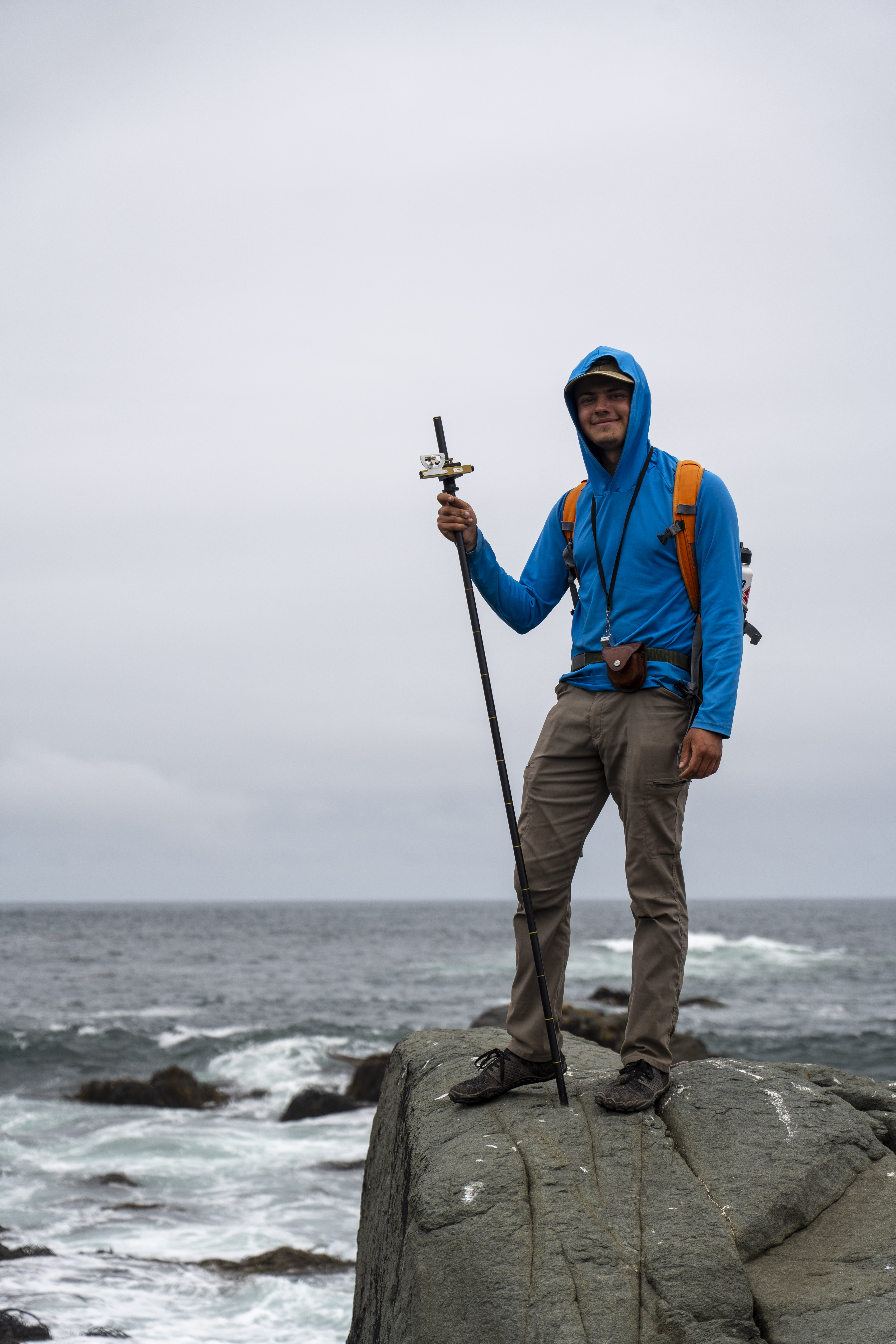 A geologist in a blue sun hoodie stands on a rocky ocean outcrop, holding a tall Jacobs staff against a backdrop of crashing waves and a cloudy sky.