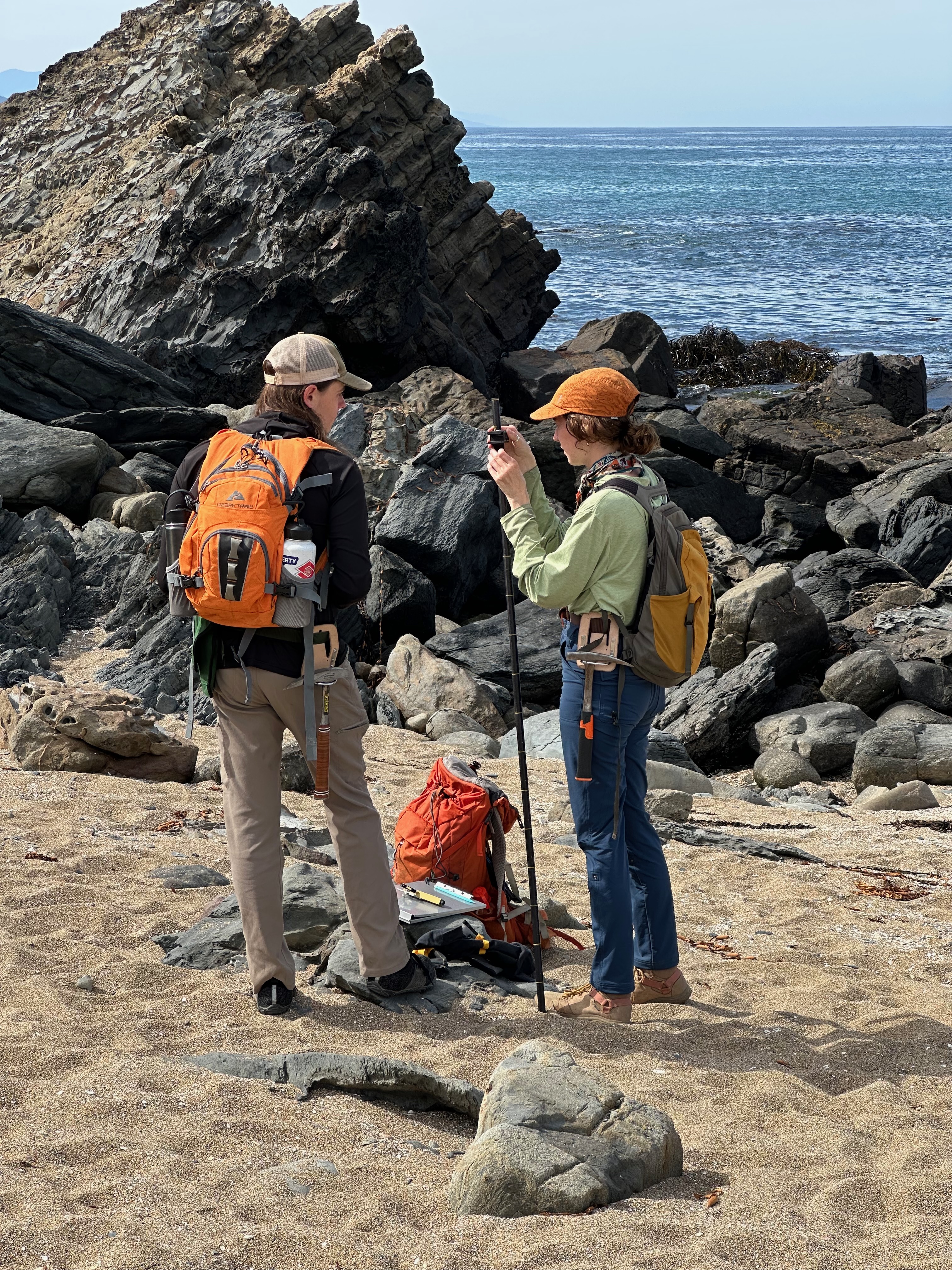 Two female geologists in hiking gear use a compass and a tall Jacob’s staff on a rugged, sun-drenched beach.