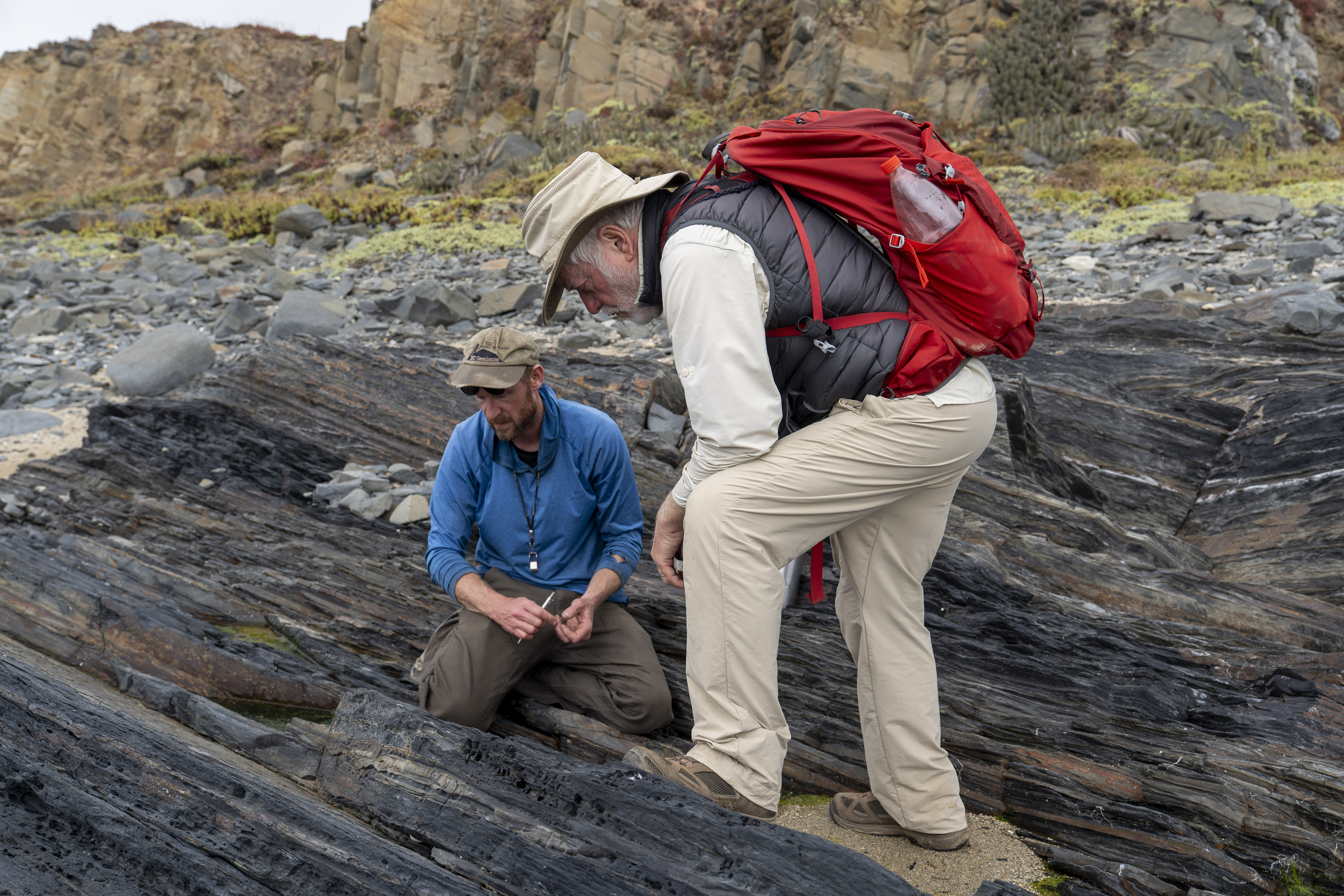 Two geologists in hiking gear examine dark, stratified rock formations on a rugged coastline.