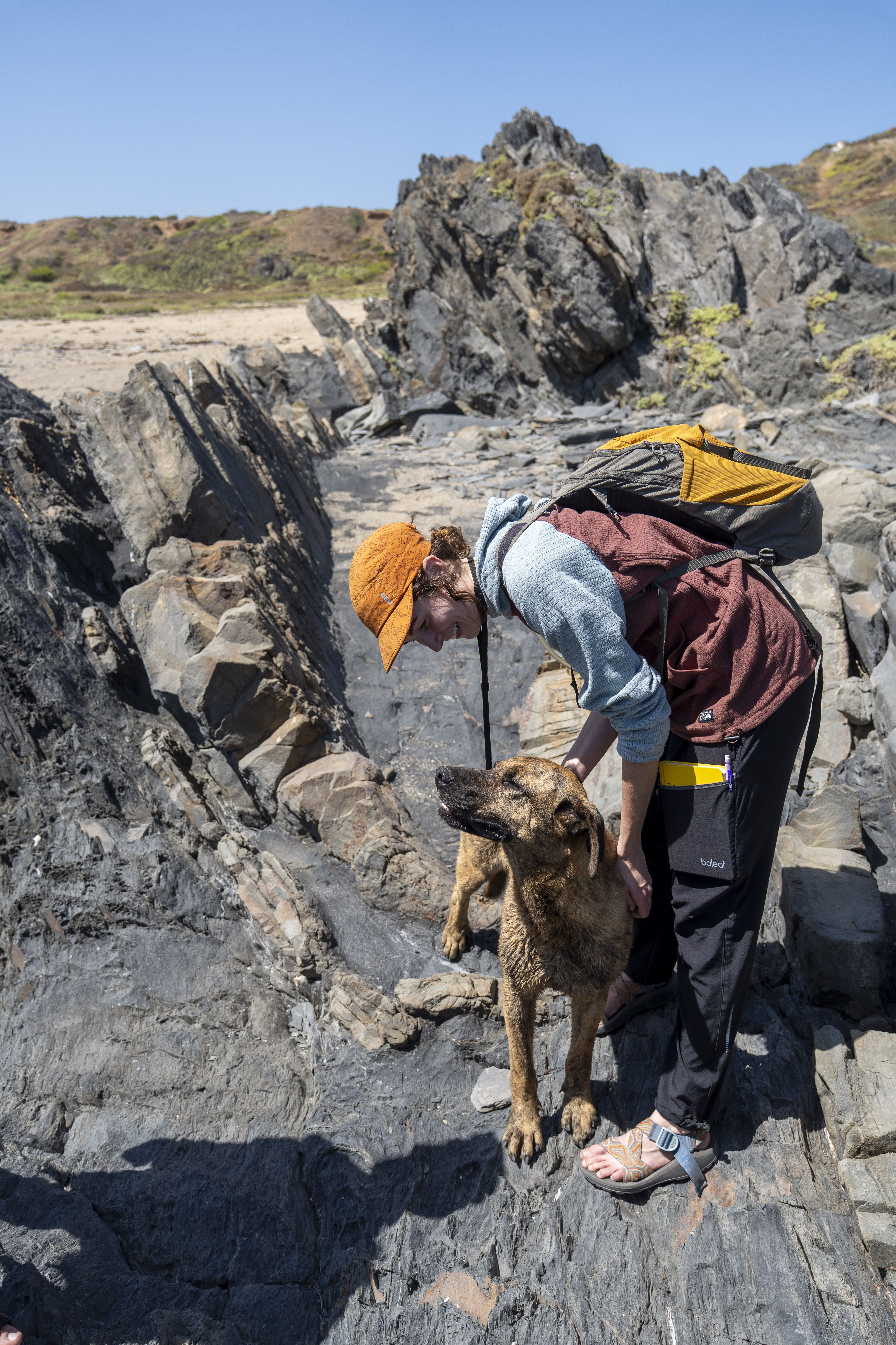 A geologist in an orange cap and backpack smiles while petting a brindle dog among rugged, dark rock formations on a sunny beach.