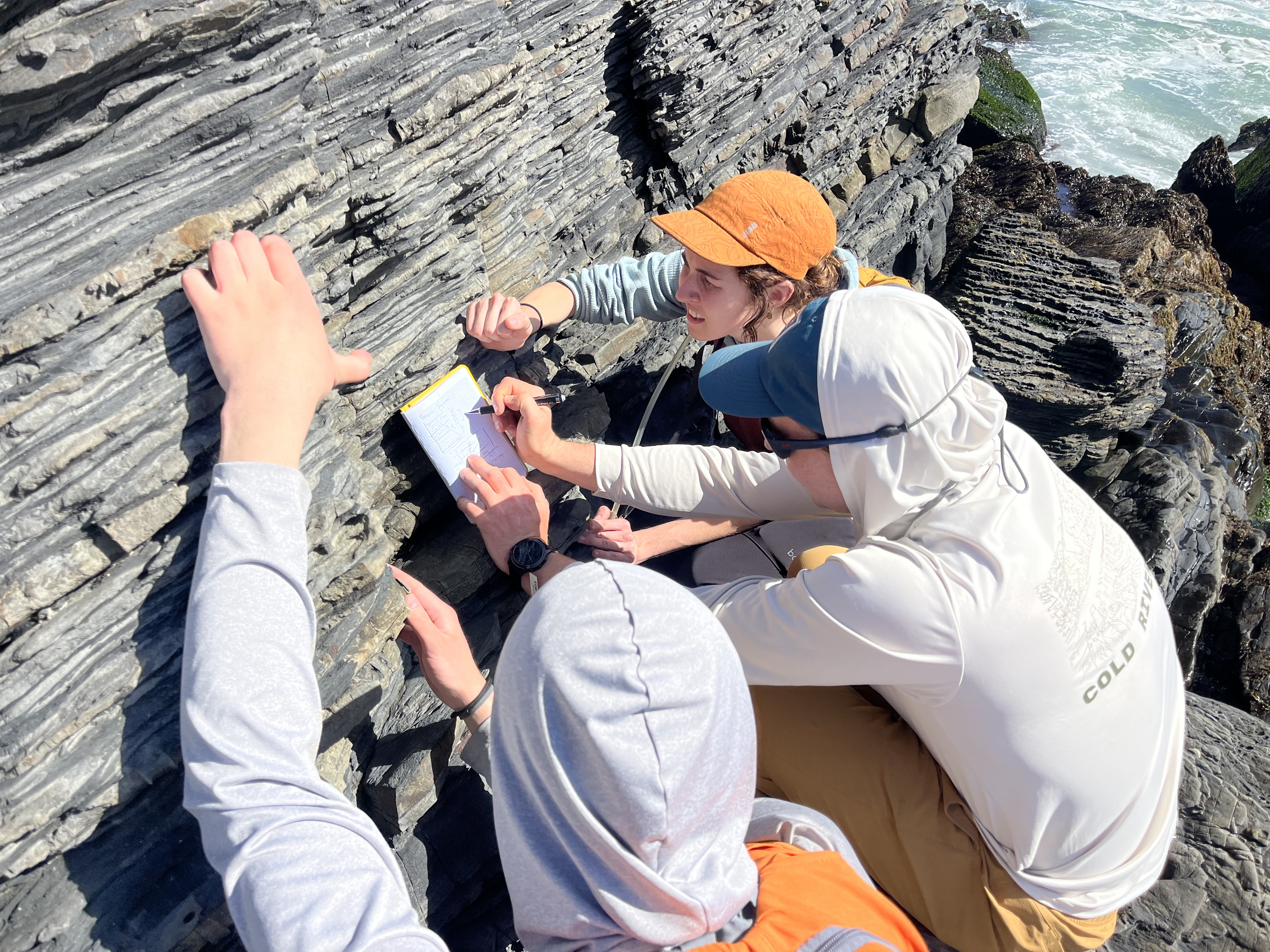 Three people examine layered rock formations by the ocean, with one taking notes while others point at the strata.