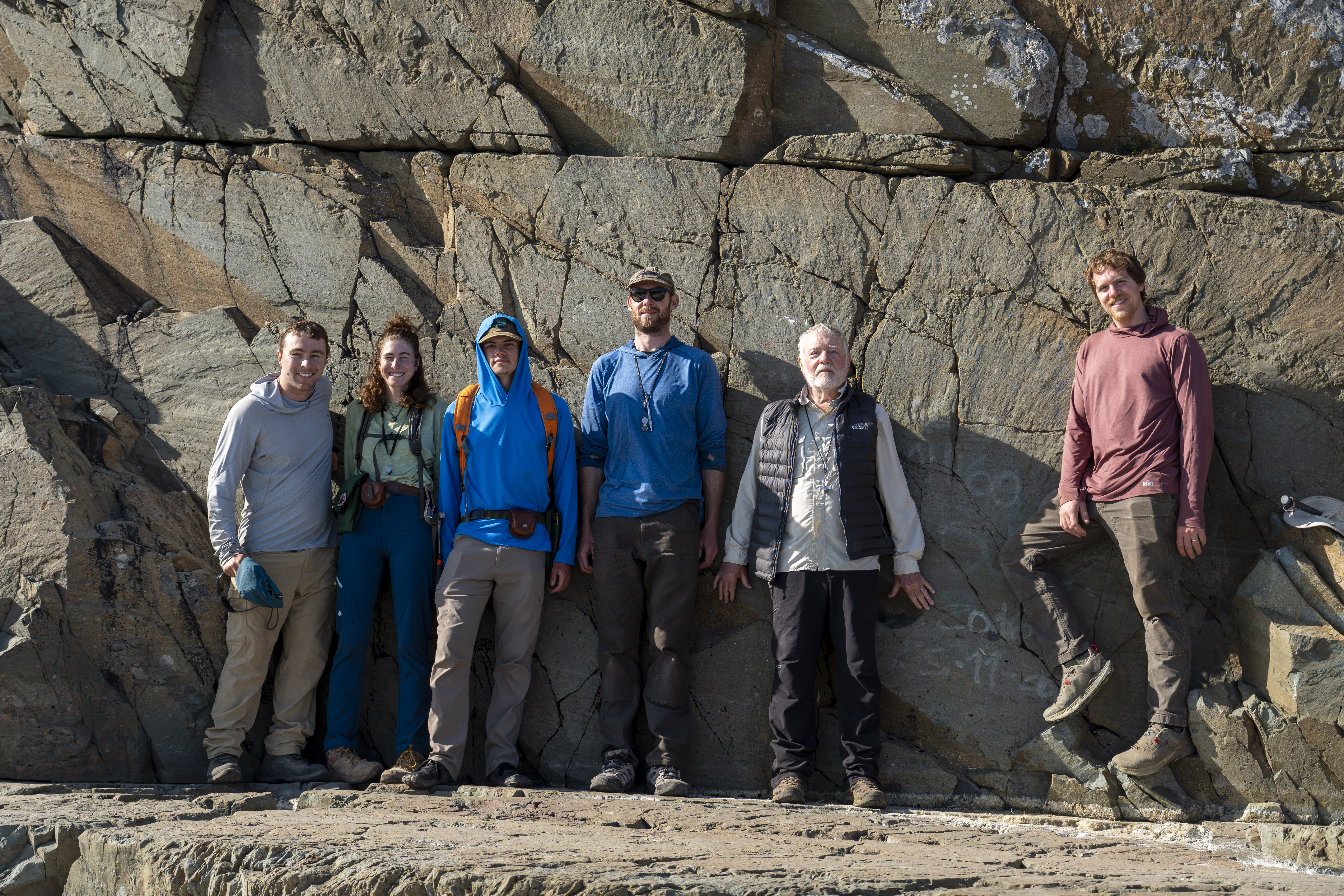 A group of six geologists in hiking gear posing in front of a massive, fractured rock wall at a sun-drenched geological site.