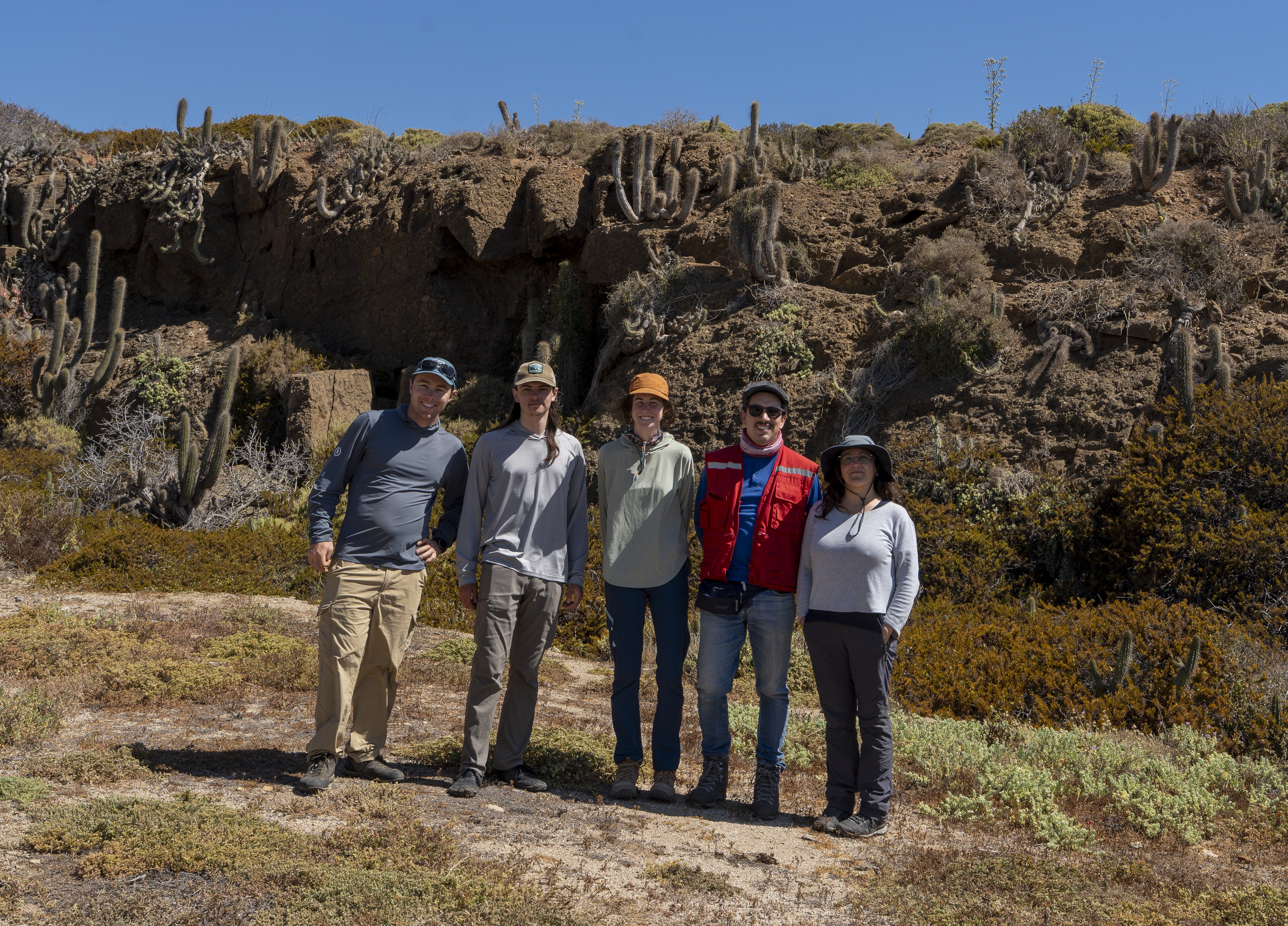 A group of five geologists in outdoor gear standing in front of a sun-drenched, rocky hillside with cacti and desert vegetation.