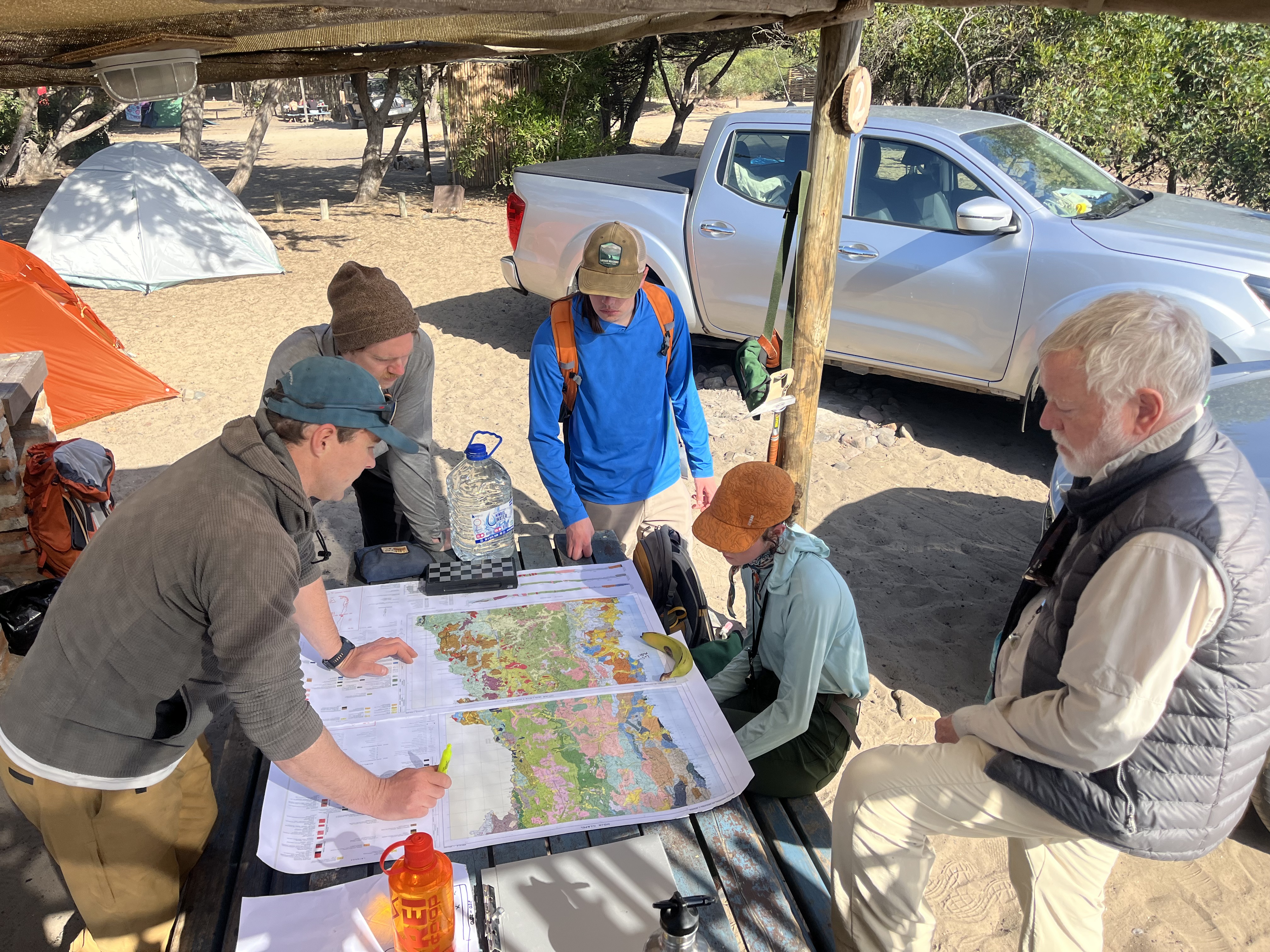 A group of five geologists and geologists in outdoor gear gather around a wooden picnic table to study large, colorful geological maps at a sun-drenched desert campsite.