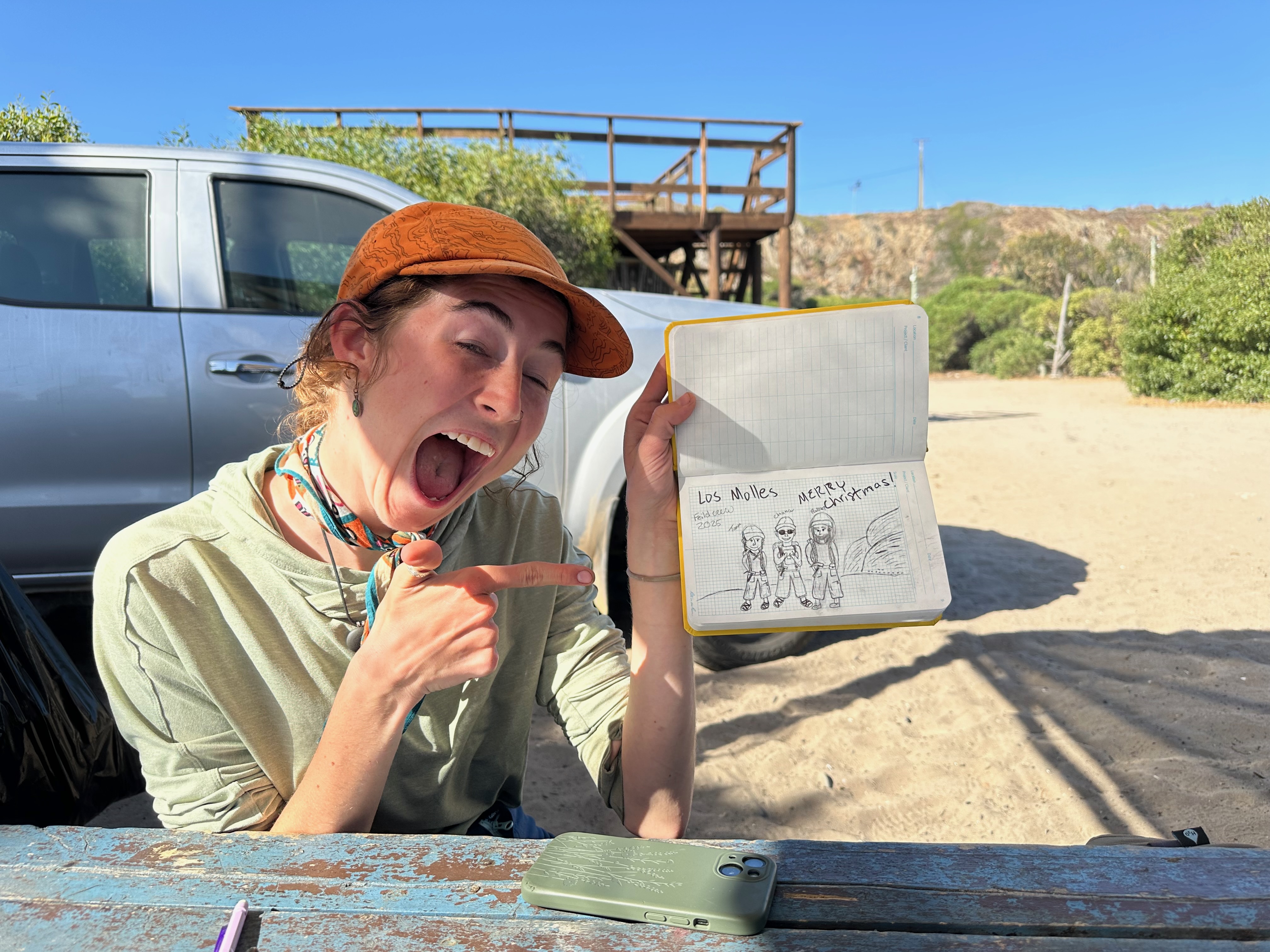 A smiling geologist in an orange cap excitedly points to a personalized Christmas sketch in her yellow field notebook at a sun-drenched campsite.