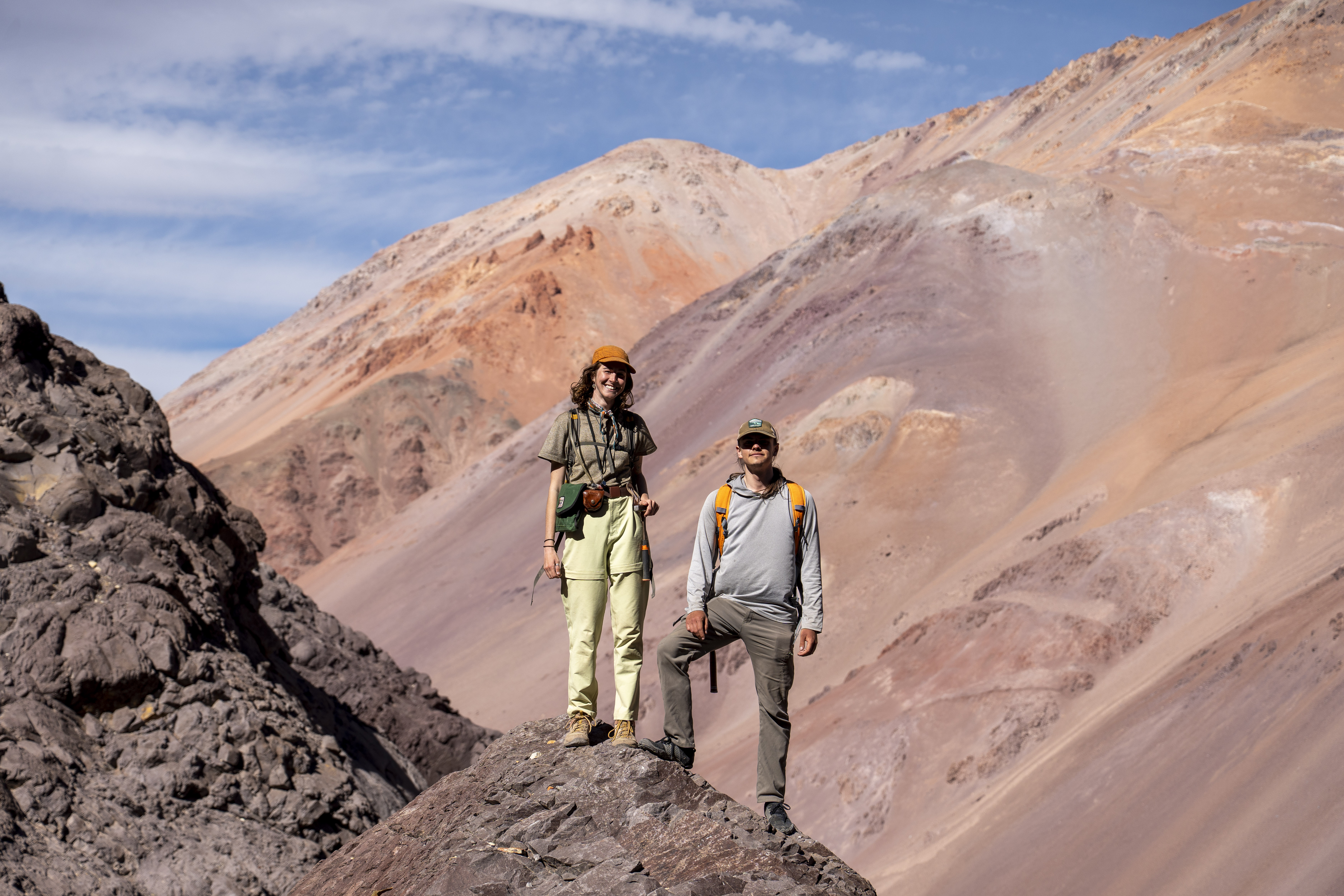 Two geologists stand on a rocky outcrop in a colorful, arid mountain landscape, posing with backpacks under a partly cloudy sky.
