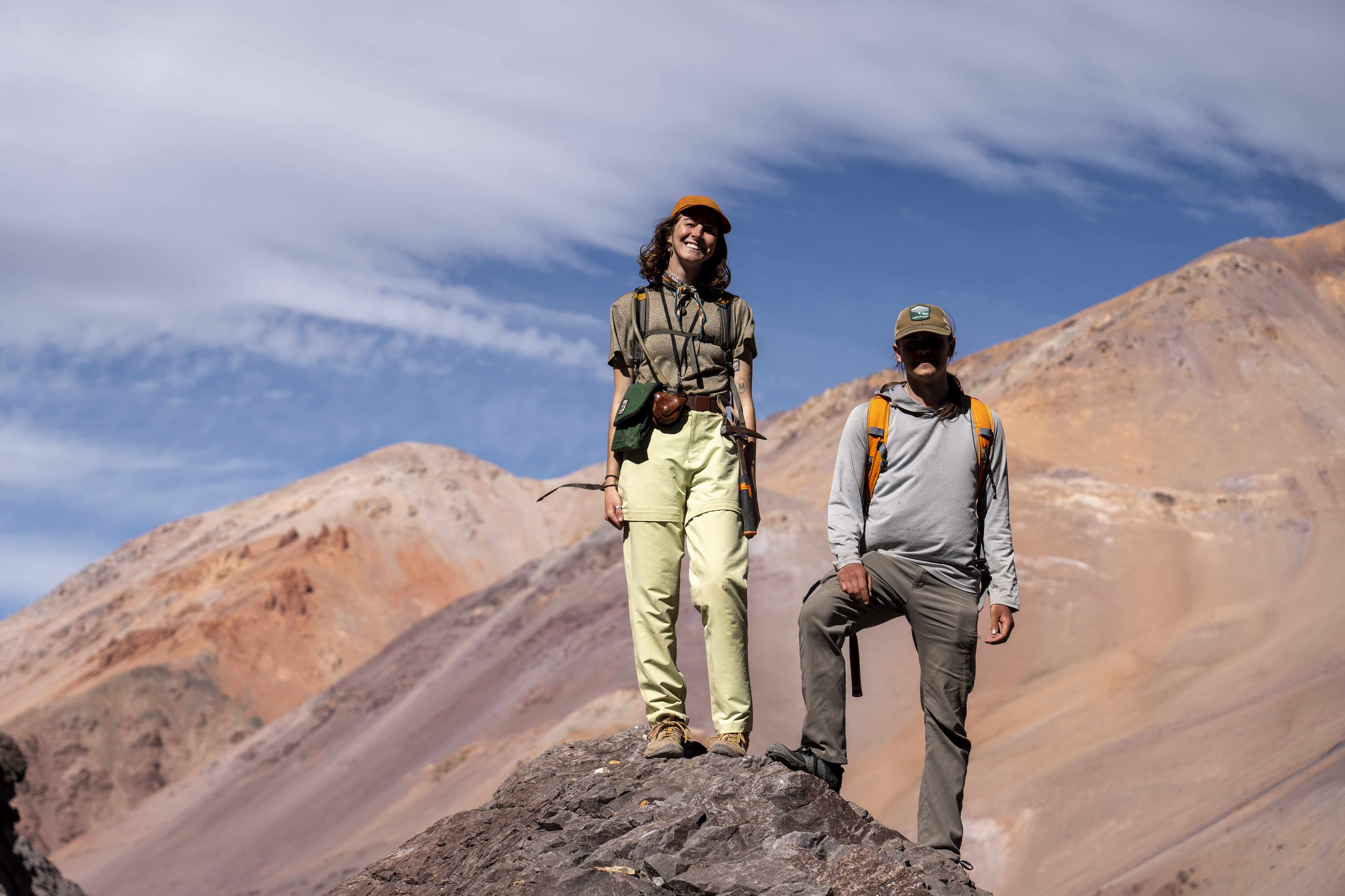 Two geologists standing atop a rocky peak against a backdrop of vast, reddish-brown mountains and a clear blue sky.