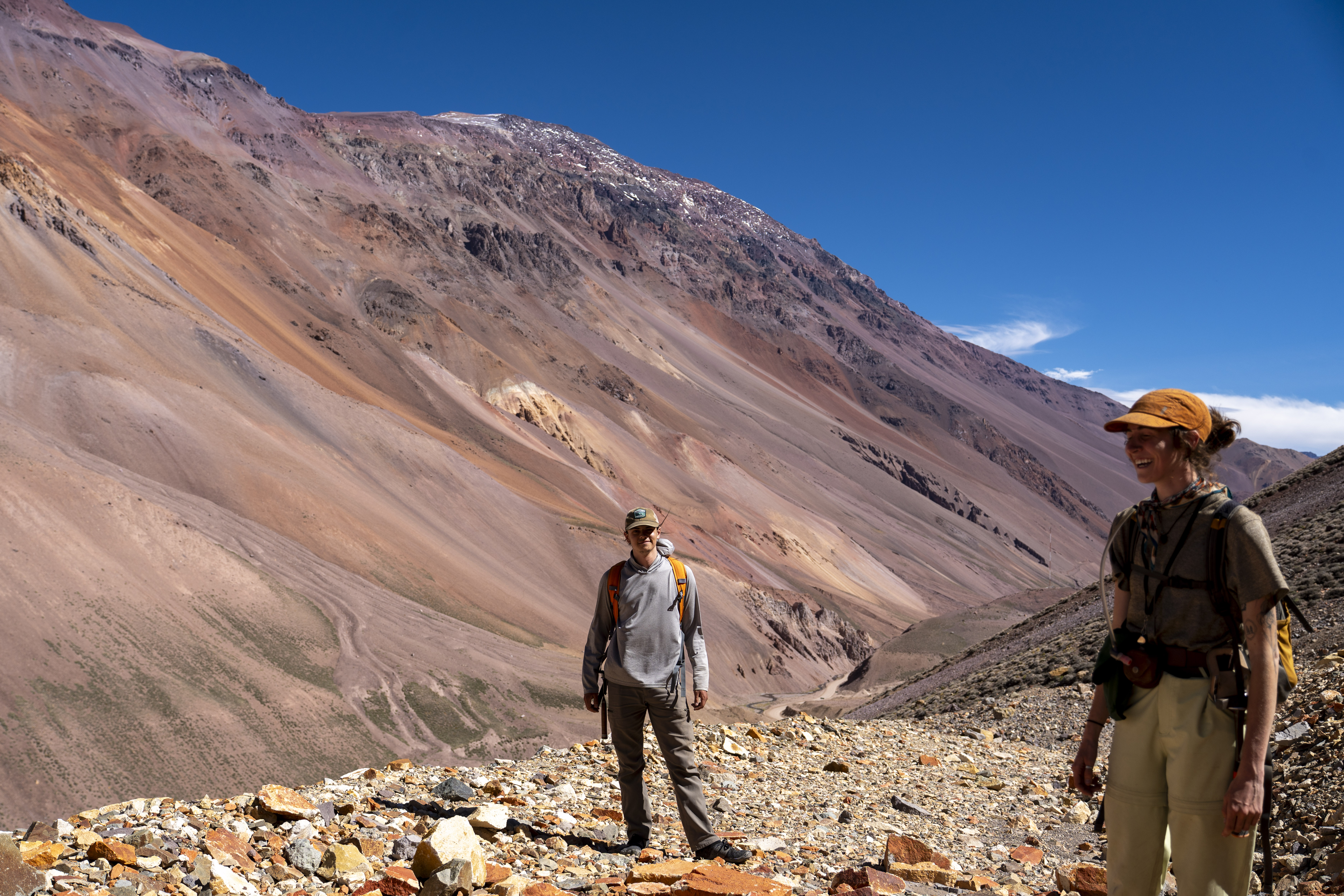 Two geologists standing on a rocky trail with vast, reddish-brown slopes and a clear blue sky in the background.