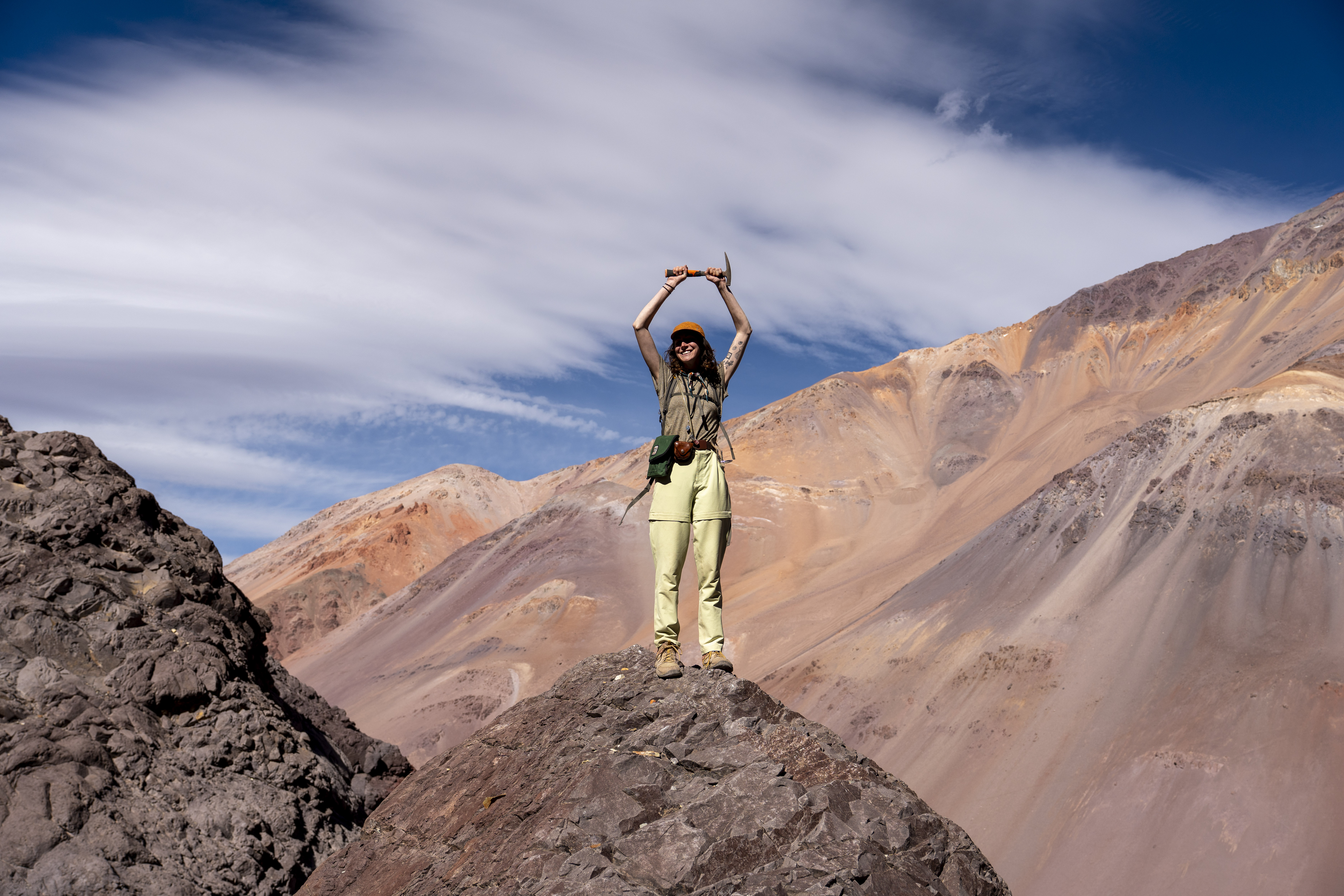 A female geologist stands triumphantly on a rocky summit, raising an rock hammer against the backdrop of the rugged, reddish-brown peaks of the Andes mountains.
