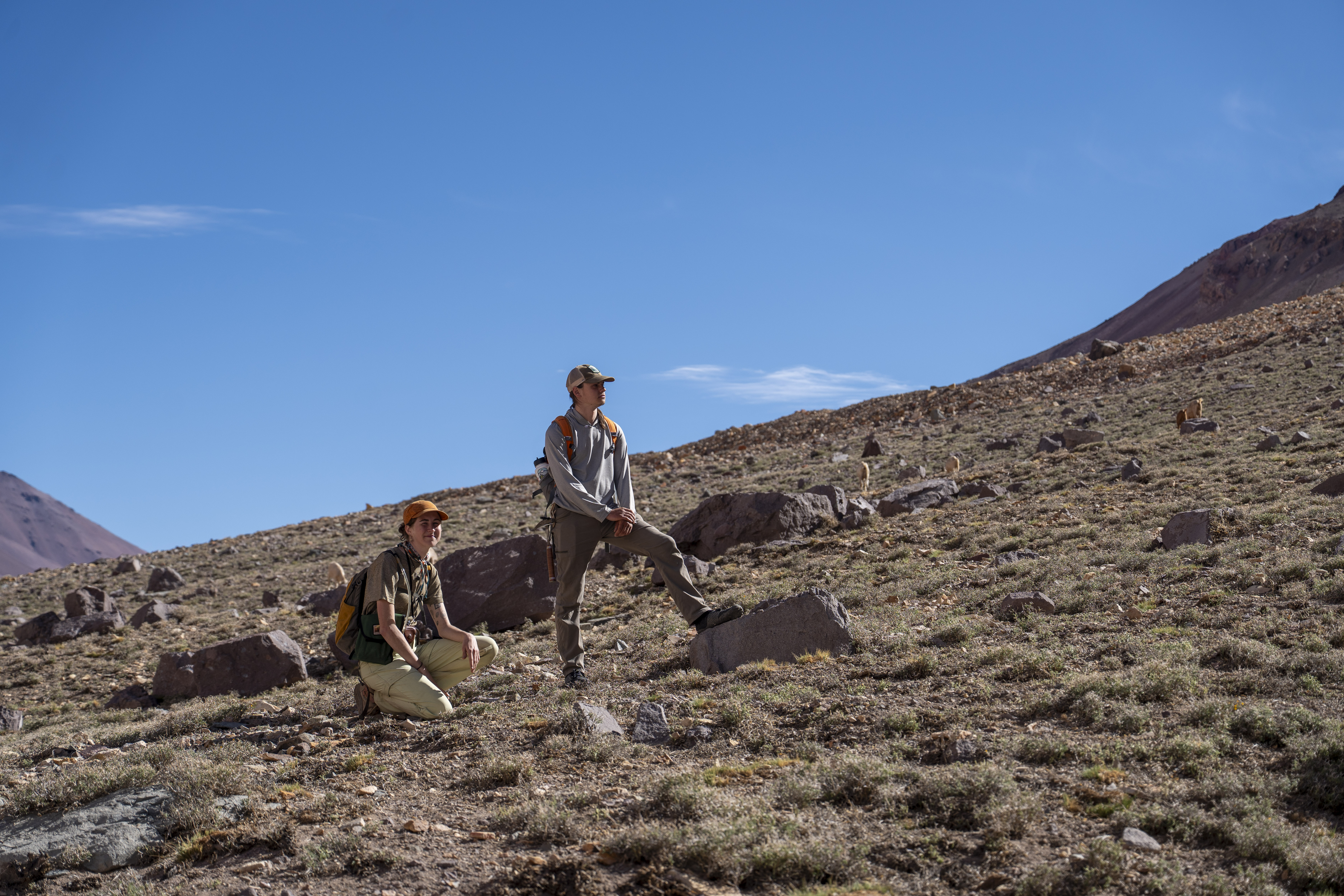 Two geologists, one kneeling and one standing, taking a break on a rocky slope in an arid landscape under a clear blue sky.