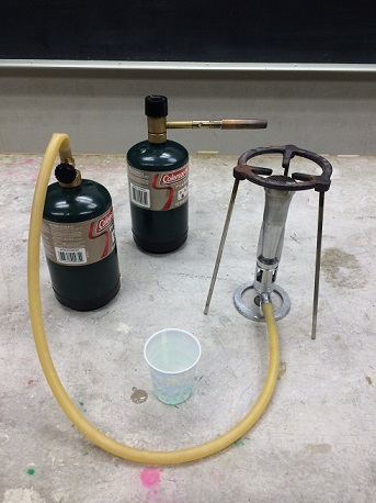 A bunsen burner by a cup and a 16 oz propane tank