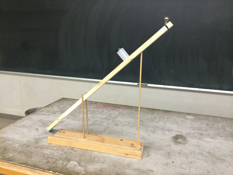 A mechanism with a stick at a 45 degree angle. The stick has a cup in the middle of it and a ball on the other end.