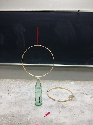 A pen sitting on top of a embroidery hoop which is on top of an old coke glass bottle