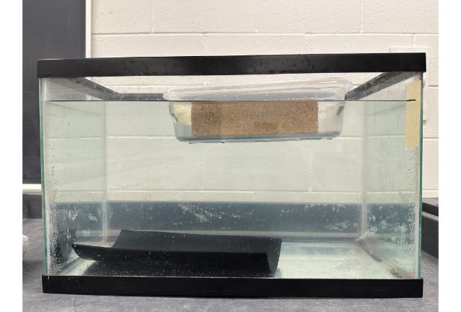 A brick floating in a clear tupperware boat in a fish tank.