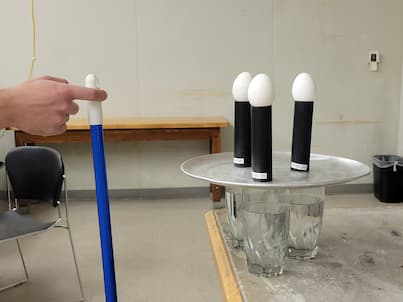 3 eggs on tubes on a pizza pan above 3 glasses of water. With a broom ready to hit the pan.