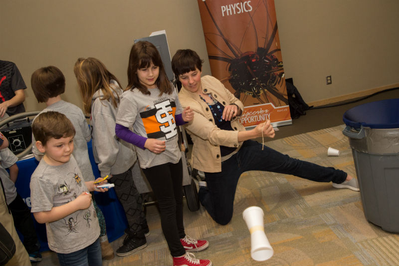 A woman and a little girl wearing an ISU shirt looking at a cup in the air as it spins from a demonstration, a sign in the background says Physics