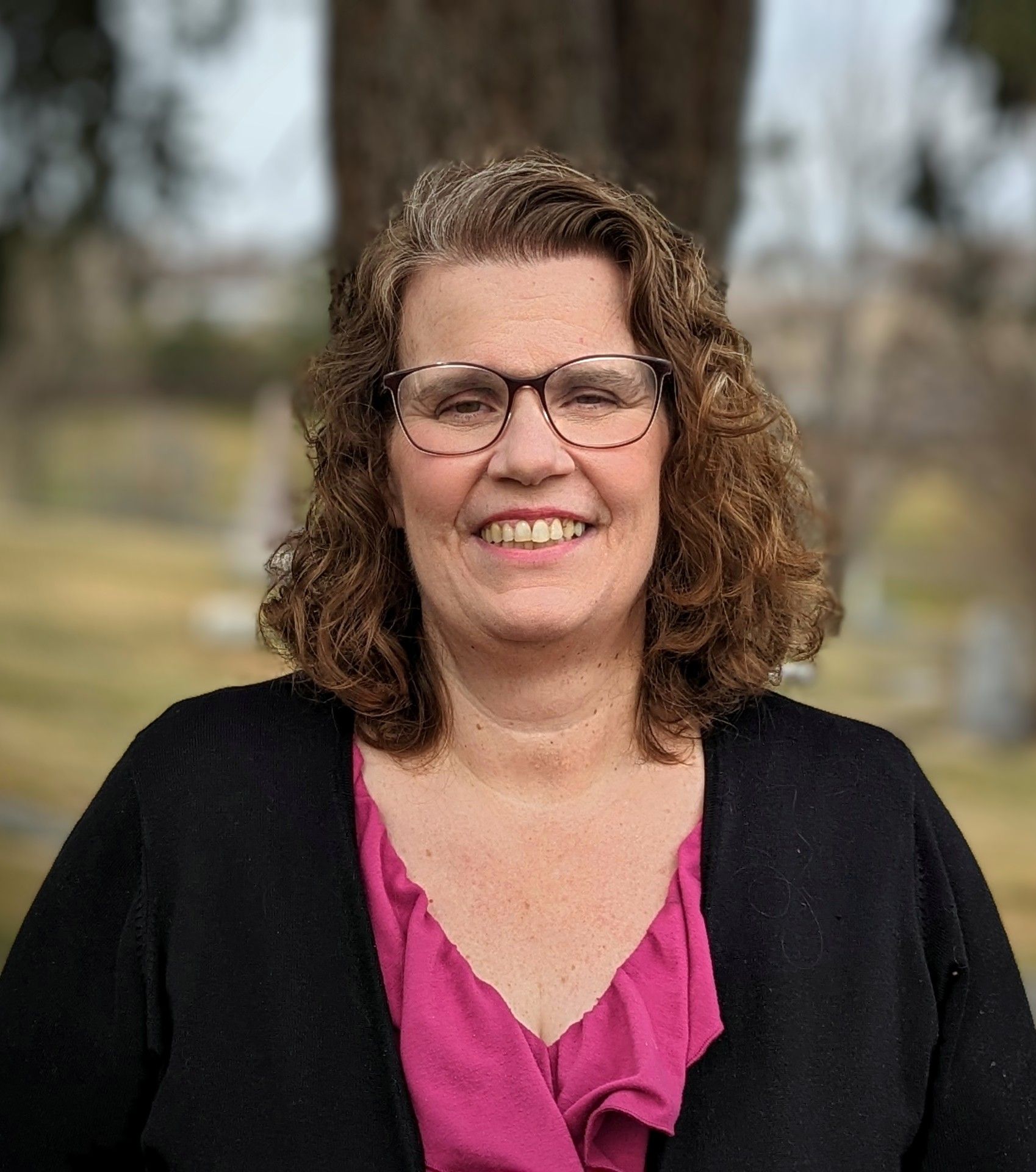 Elizabeth Schniedewind, a white woman in her 50s is pictured wearing a black jacket with a pink dress. She wears glasses and has brown curly shoulder length hair, standing in front of a tree