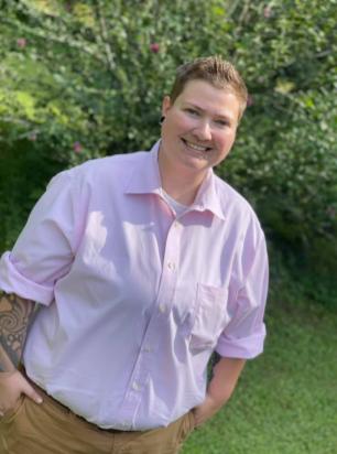 Nicole, a white-presenting female is wearing a pink button-down shirt, has short brown hair, a black earlobe plug and two silver stud piercings below her bright smile.