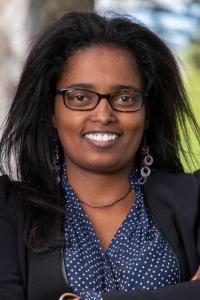 A woman with dark skin tone. She has black hair, tortoiseshell glasses and brown eyes. She is a blue button down shirt and a black suit jacket. 