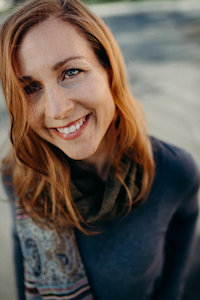 A woman with a light skin tone and red hair. She is wearing a long sleeved grey hair and a patterned scarf. She is smiling.