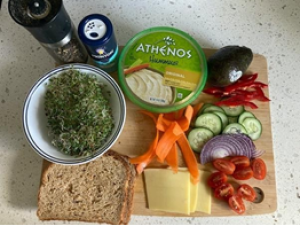 wheat bread, sliced cucumbers, carrot ribbons, sliced red onion pepper grinder, sliced cherry tomatoes, container of hummus, 2 slices of cheese, avocado. salt shaker, alfalfa sprouts, sliced red bell pepper- all sitting on a wooden cutting board