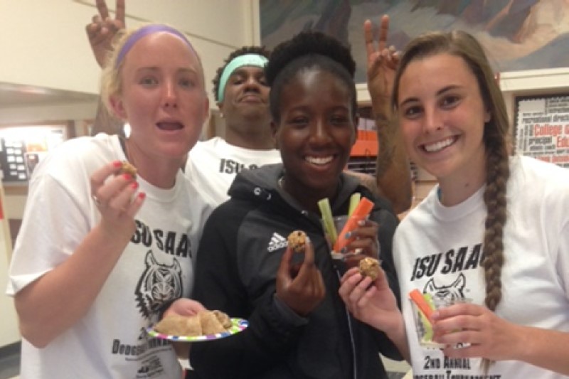 Three ISU female athletes (one African American, two caucasian) enjoy a healthy snack, a fellow male athlete looks on 