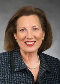 Mary Nies headshot with blue and gray plaid jacket, dark brown hair, pearl earrings. 