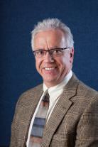 Mark Roberts professional headshot in glasses, tan suit jacket, blue background