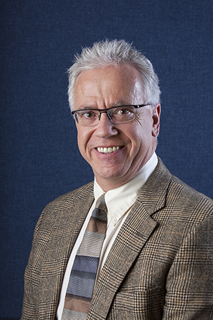 Mark Roberts professional headshot in glasses, tan suit jacket, blue background