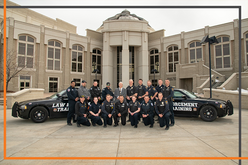 Sixteen police officers and two police cars pose in front of Stephens Performing Arts Center