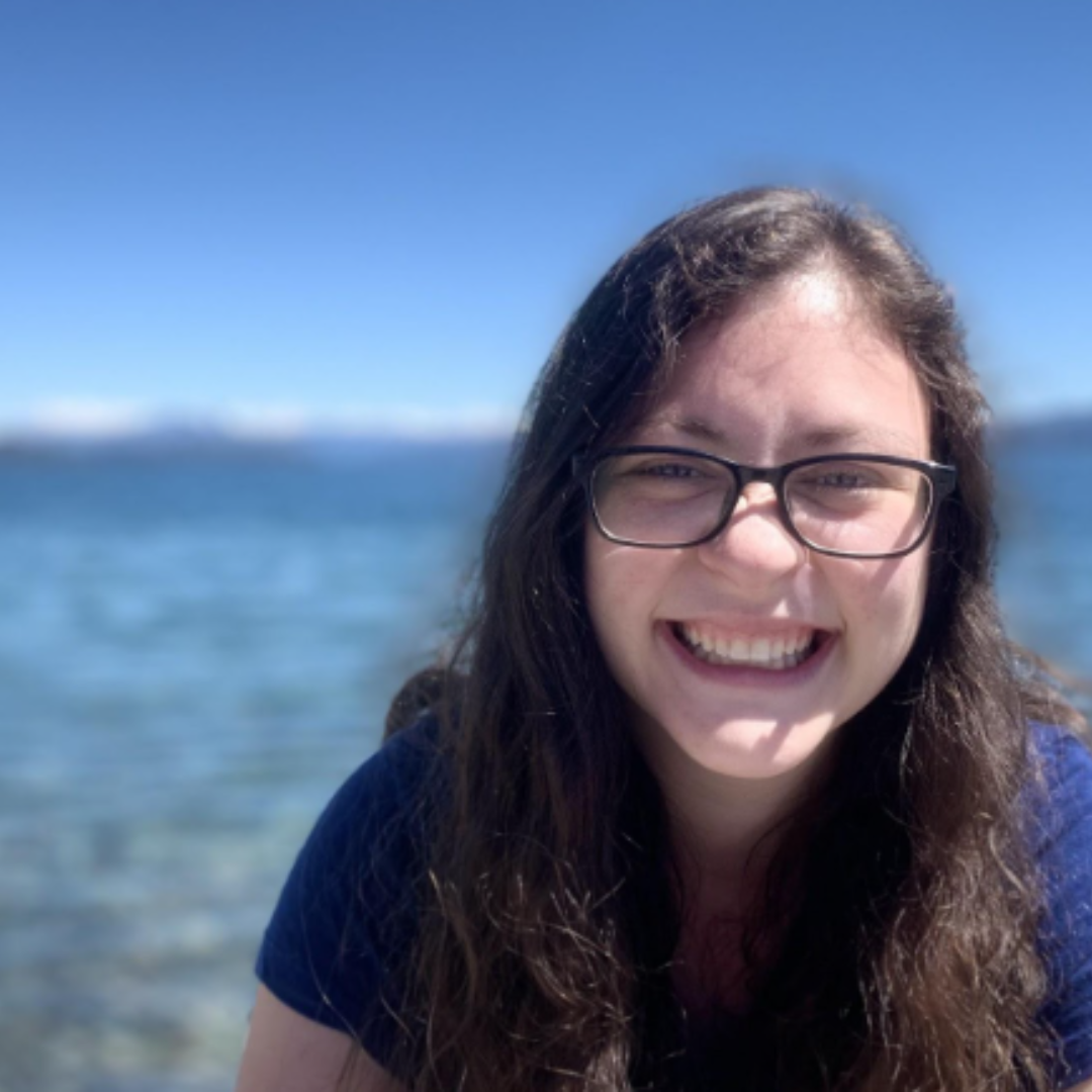 headshot of Jess standing in front of blue sky and blue water