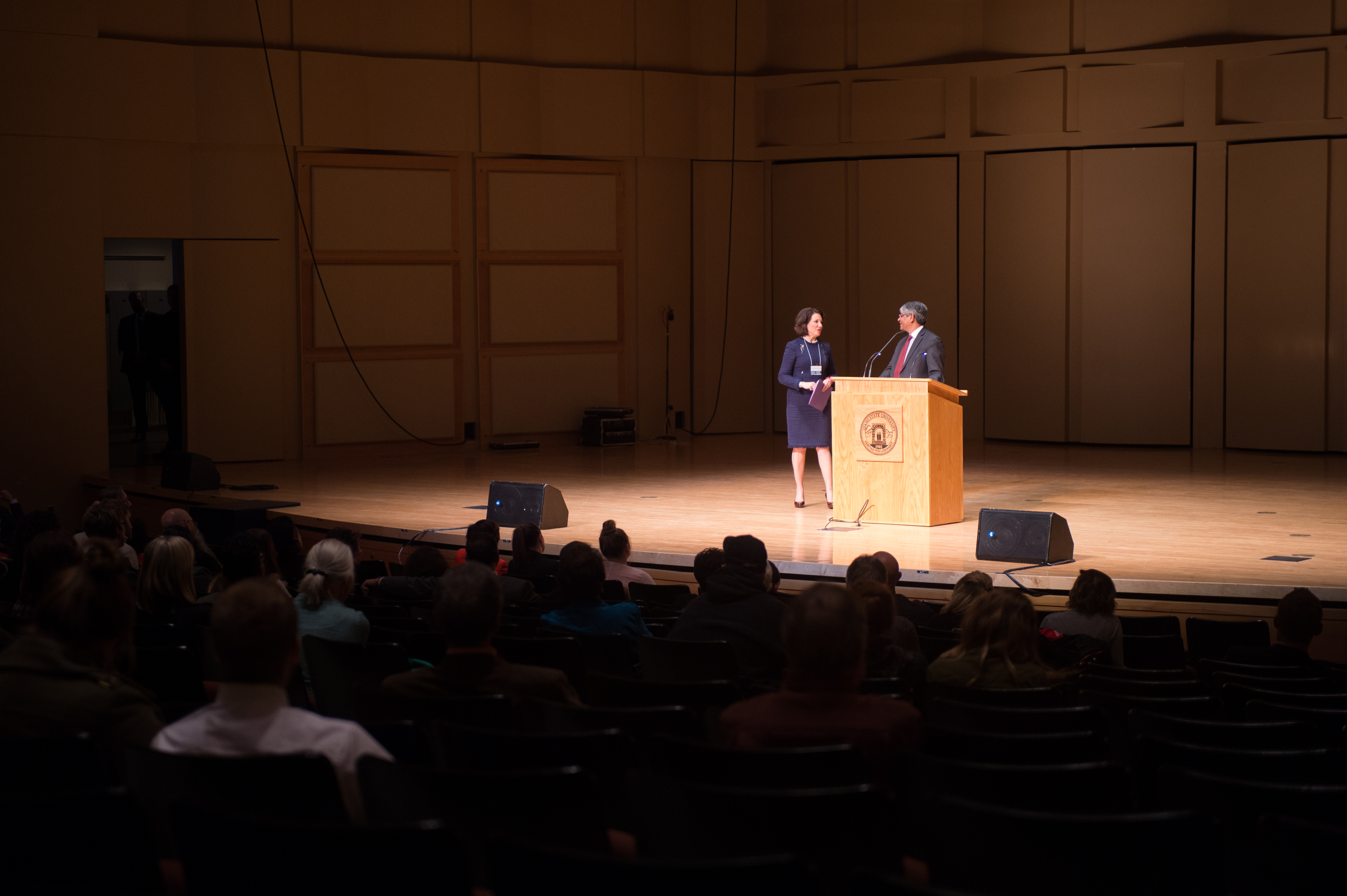 Two speakers standing on stage in a full, dimly lit auditorium.
