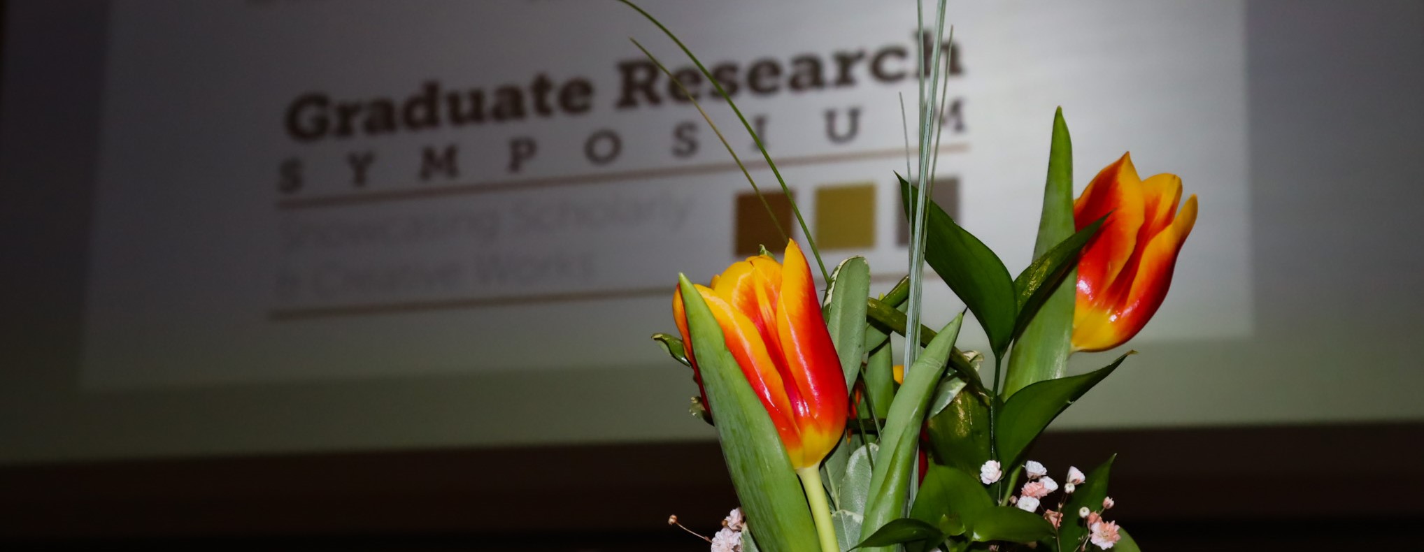 A vase of flowers sitting in front of the Graduate Research Symposium Logo on a screen
