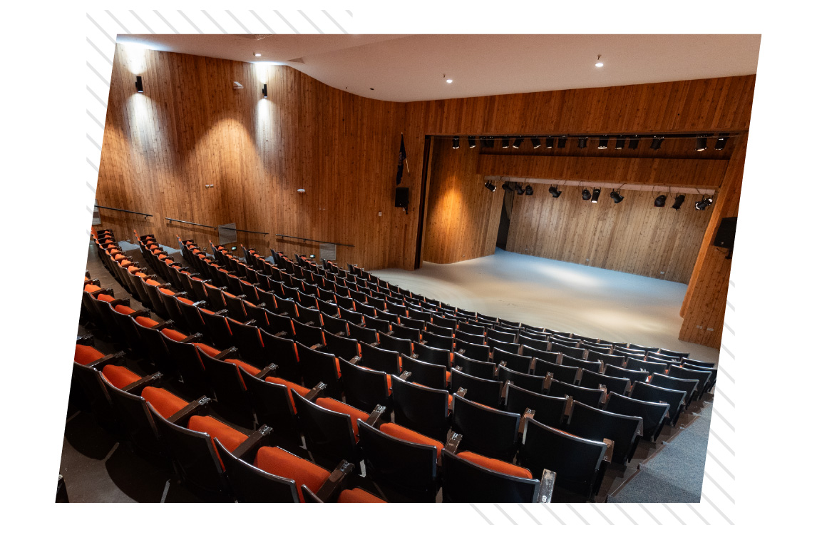 Angled interior view of a large auditorium with tiered rows of black chairs featuring orange seats, facing a wide wooden stage with overhead stage lighting; wood-paneled walls and ceiling lights create a warm, professional performance and lecture setting.