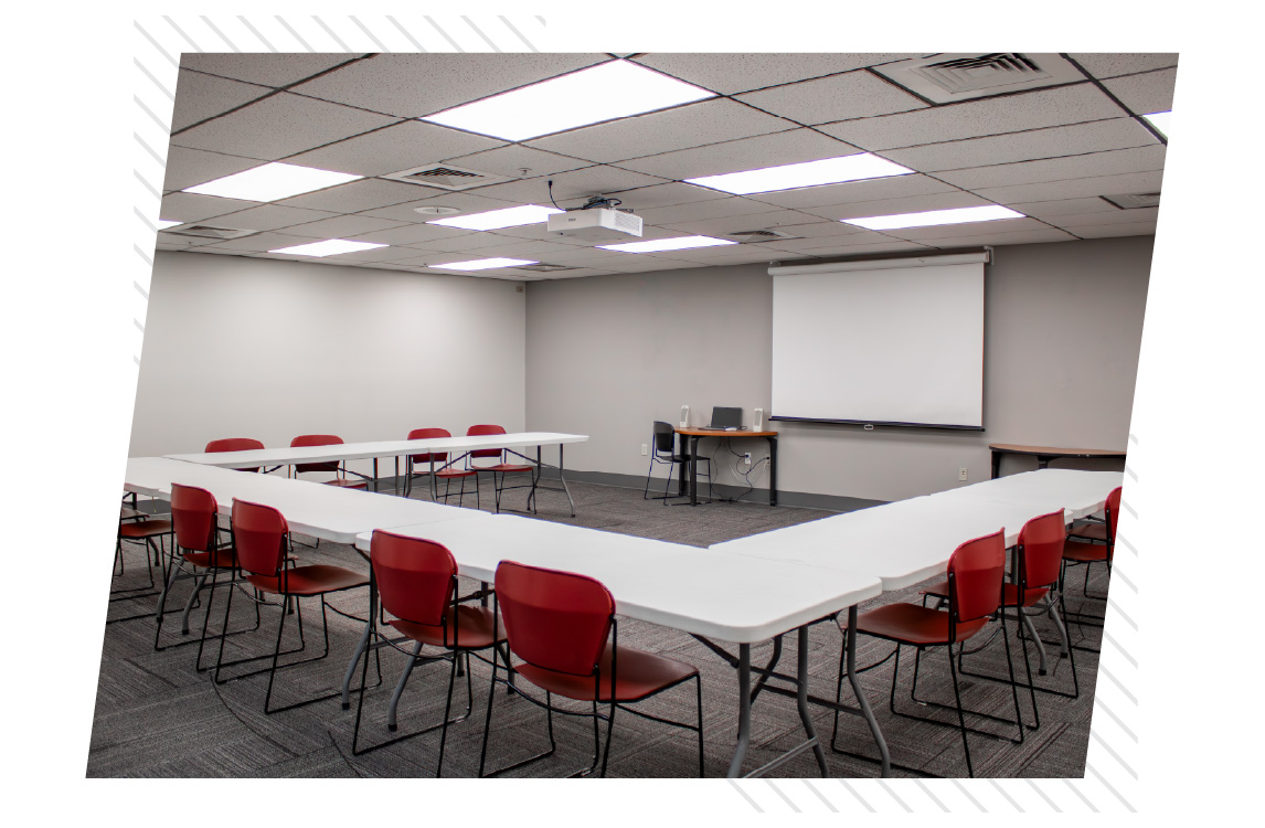 Meeting room arranged in a U-shape layout with white tables and red chairs, facing a ceiling-mounted projector and pull-down screen with a computer on a front table.