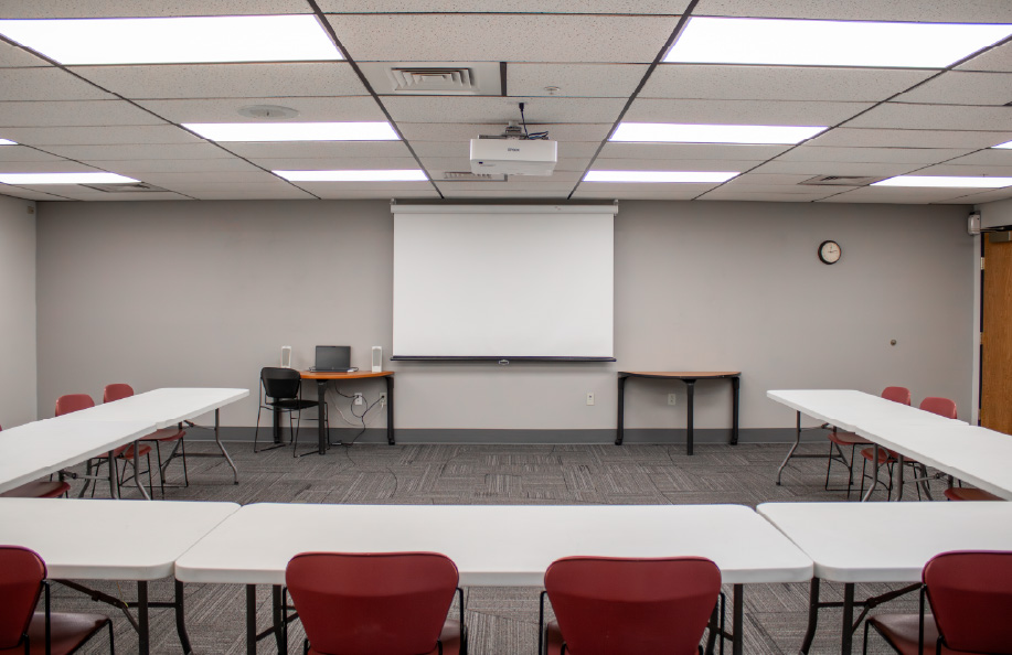 Meeting room arranged in a U-shape layout with white tables and red chairs, facing a ceiling-mounted projector and pull-down screen, with a laptop and speakers on a front table.