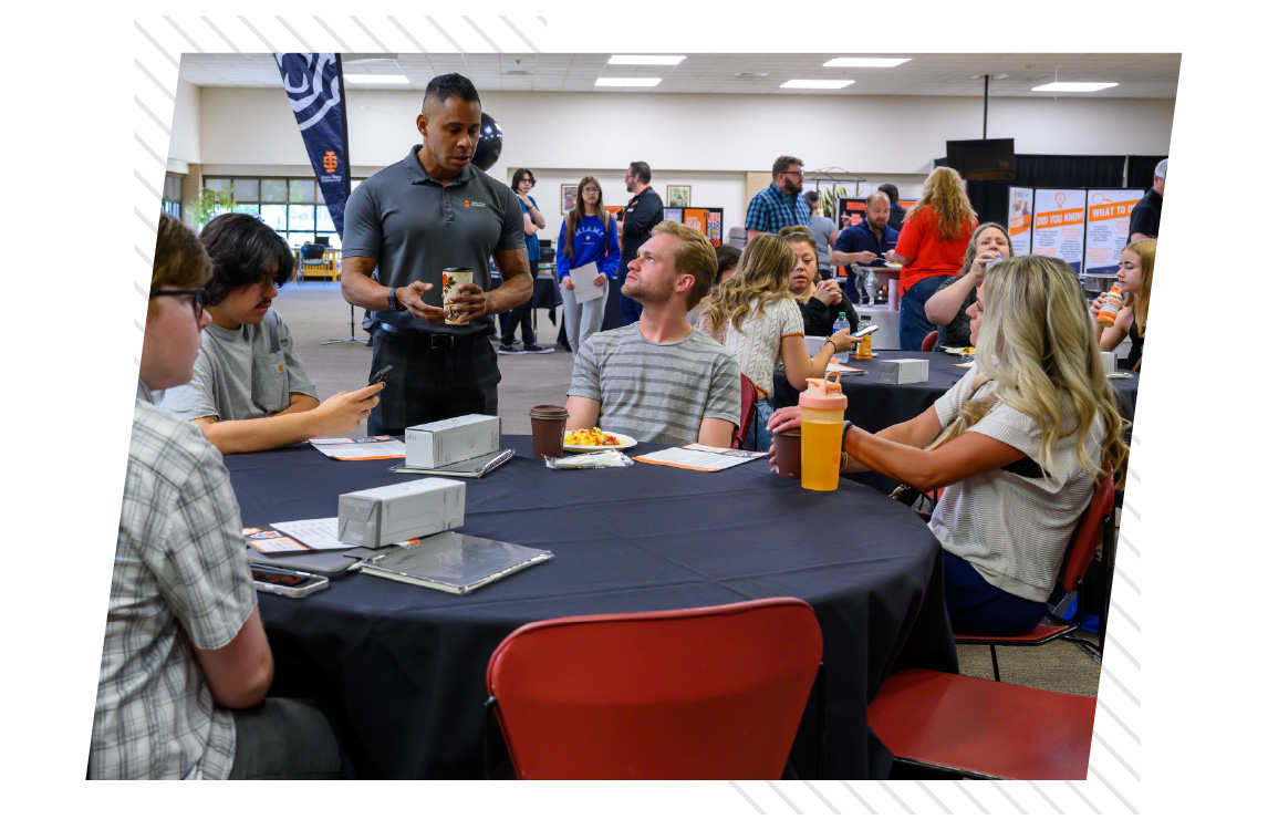 Group of students and staff seated around round tables during a campus event, talking and sharing food in a large indoor space with informational displays in the background, illustrating a banquet-style setup for networking or community gatherings.